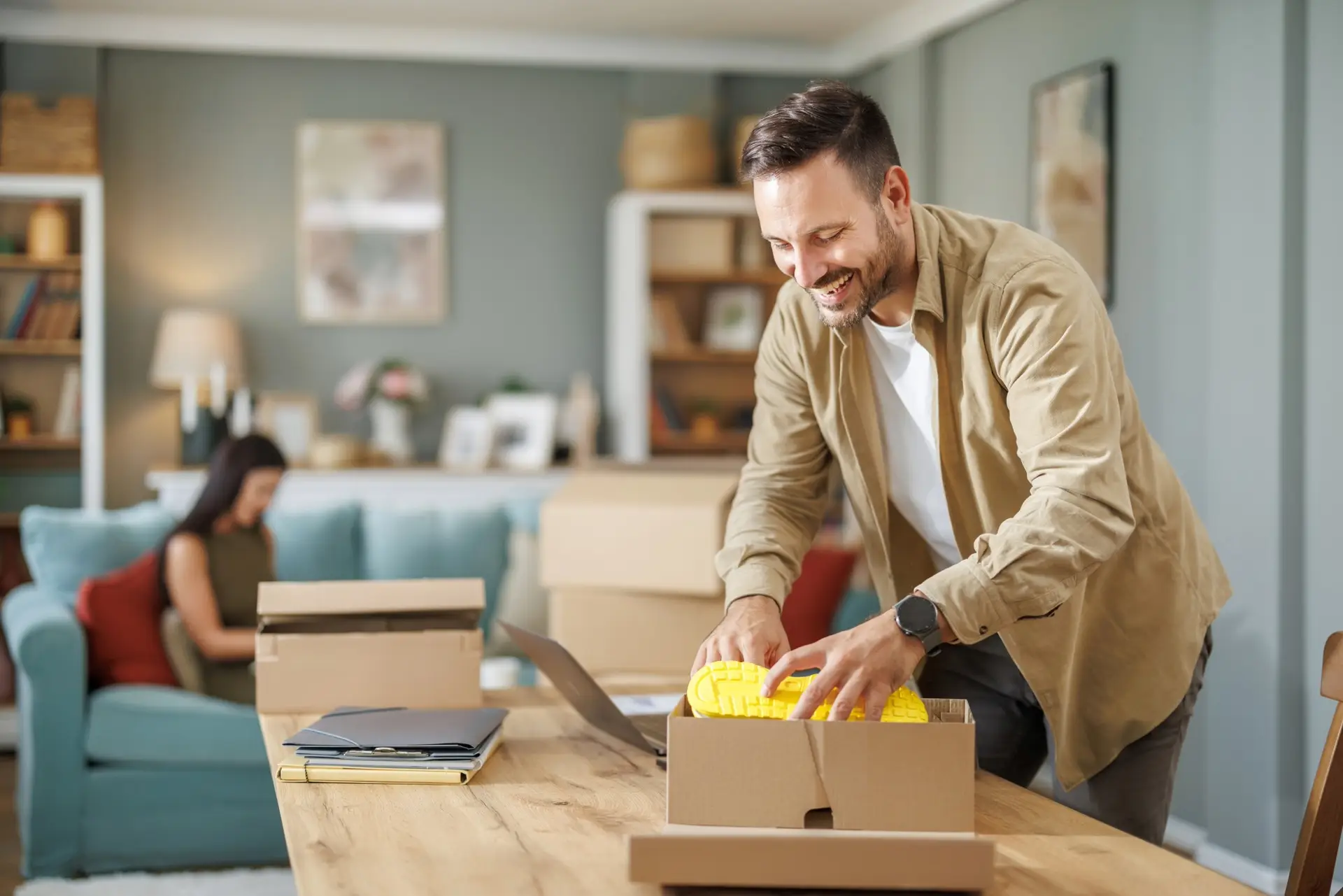 with a smile on face, a mid aged man putting new shoes in a shipping box at his nice home desk which is a simple wooden table and wooden chair
